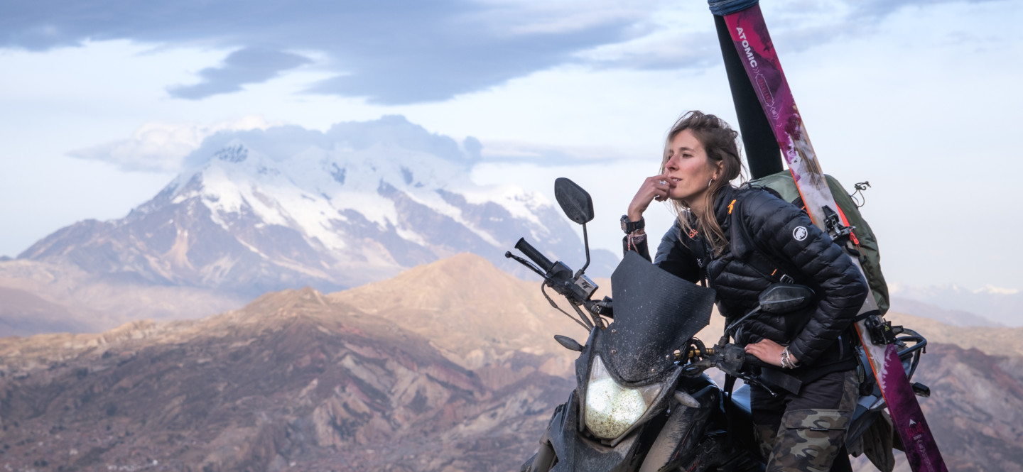 Na zdjęciu kobieta siedząca na motocyklu. Na plecach ma narty. W tle góry